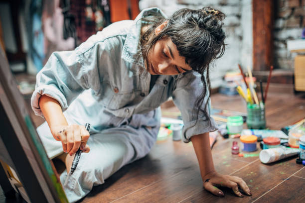A young woman is sitting on the floor of an art studio, painting on a canvas. She is wearing a jumpsuit, with her hair tied in a bun. Surrounding her are paints, brushes, and art supplies.