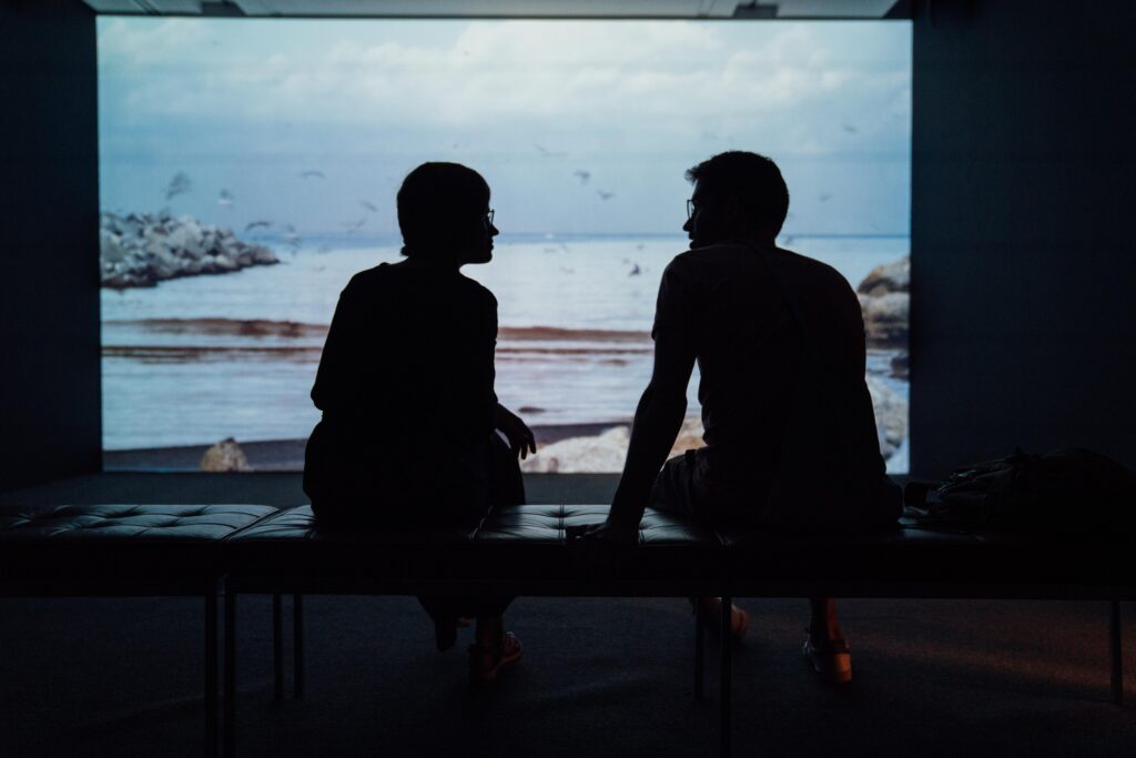 In this image, a couple is sitting next to each other in a dark room. In front of them is a projector, showing images of a beach. We see their silhouette as they look at one another.