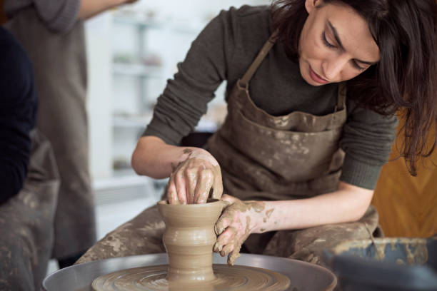 A young woman is in a pottery class, creating a piece of art on a pottery kiln. She is using her hands to shape the clay. Her brown apron is covered in clay.