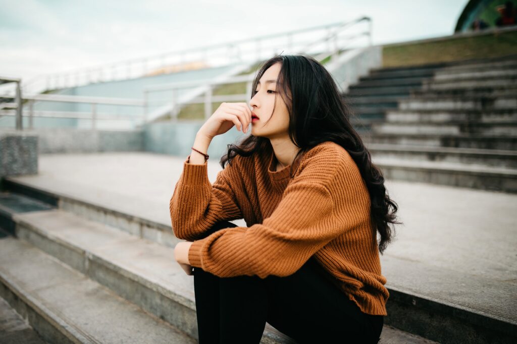 A young Asian woman is sitting on concrete steps outside as she thinks to herself. Her arm is resting on her knees, while her hand is over her face.