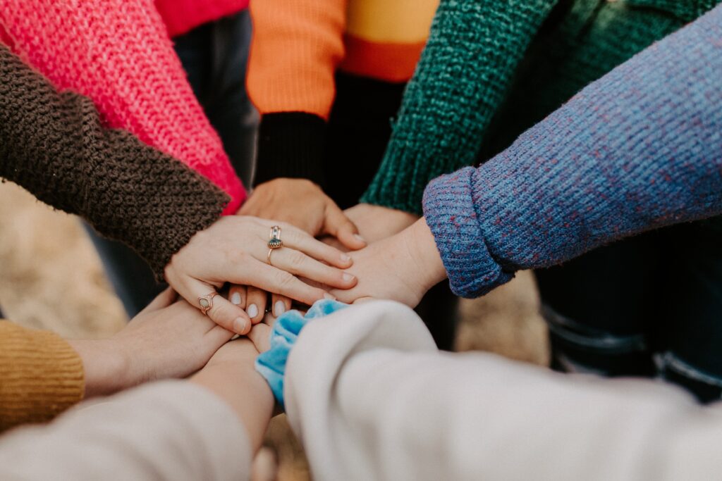 In this photo there are 8 people putting their hands into a group huddle. They are all wearing long sleeve tops and have their hands on top of each other.