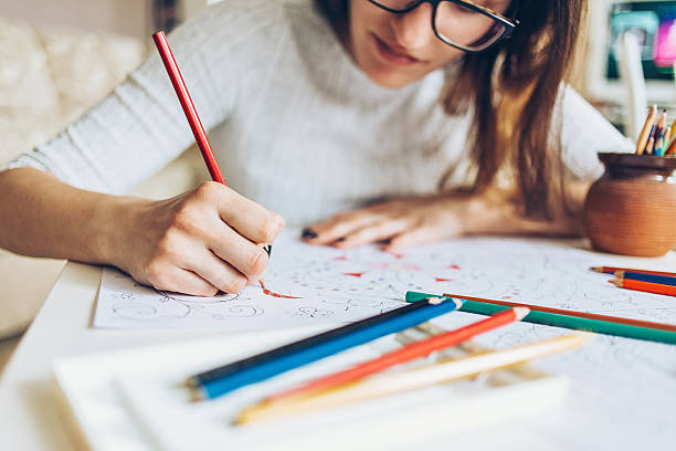 A young woman wearing reading glasses is sitting at a desk using colored pencils to draw. She is using the red colored pencil to draw on a piece of paper, as there are different colored pencils across the table in front of her.