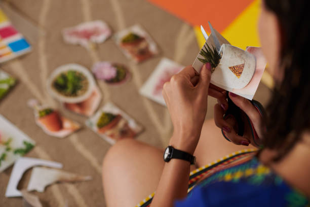 In this photo, a young woman is cutting out images of plants out of magazines using a scissor. She is cutting these images out and laying them in front of her to create a collage.