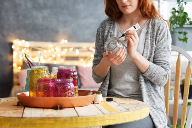 A red headed young woman is sitting at a wooden table with painted jars of different sizes on the table. She is creating worry jars and is doing arts and crafts.