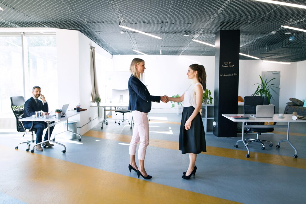Two business women are standing in an office building shaking each other's hands. A business man is sitting on the far left of the photo at his desk, glancing at the two women.