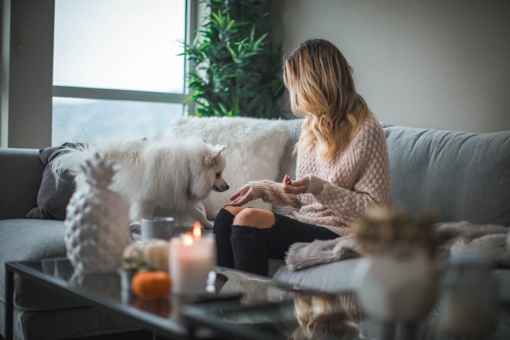 A young woman is sitting on her couch in her living room at home. Her small white dog is on the couch beside her sniffing her hand. There is a lit candle on the coffee table in front of her.