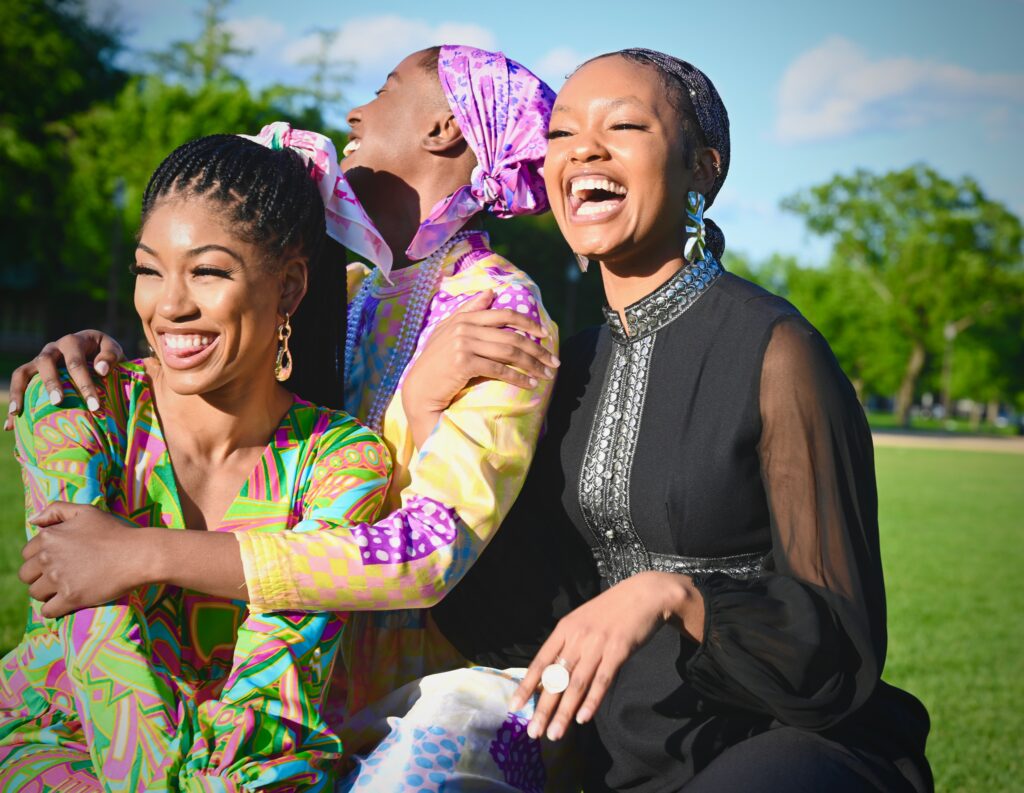 Three young African American women are at a park enjoying the outdoors. They are all embracing each other, closely seated and smiling and laughing.
