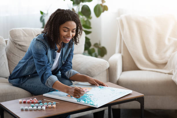 A young African American woman is sitting in her home in the living room. She is seated on the couch and has different colored paints on the coffee table, as she uses them to create a painting.