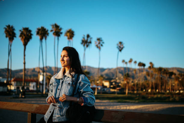 Portrait of a brunette woman, enjoying the ocean view in the magic sunset hour. She is wearing fashionable, retro styled streetwear, a denim jacket and sunglasses, under the California sunshine.