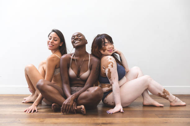 A group of 3 women are sitting on the floor posing for their photo to be taken. They are all wearing lingerie. The woman on the left is a Hispanic woman smiling, in the middle is an African American woman, and on the right is an Asian American woman with vitiligo.