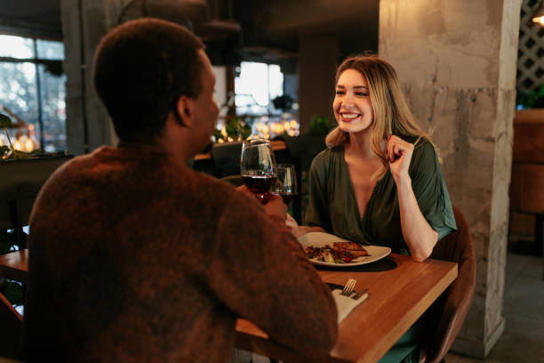 A young interracial couple is at a date at a restaurant sitting across from each other. The man is facing the camera backwards, as the woman sits in front of him smiling.