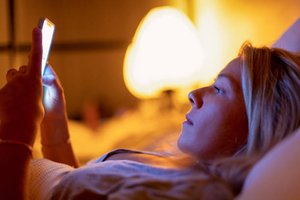 A blonde young woman is laying in her bed at night looking at her phone. She has a serious facial expression as the light from her phone shines on her face.
