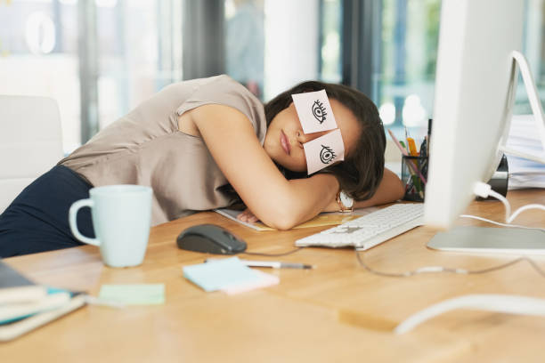 A young businesswoman is at her office, taking a nap at her desk. There are sticky notes covering her eyes, and they each have a doodle of an eye on them. Her desk is covered with a coffee cup, pens, and sticky notes.