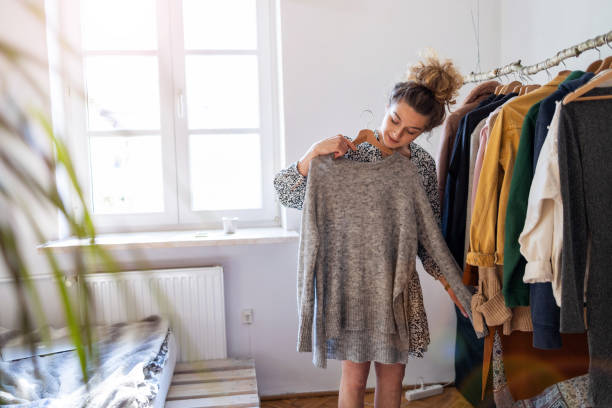 A young woman is standing in her closet trying on clothes. A rack of clothing is hanging next to her as she holds up a dress against her.