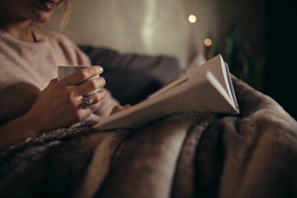 In this image, there is a cropped image of a woman sitting on her couch, covered in a blanket. On her lap there is an open book and she is holding a cup of tea in her hand as she reads.