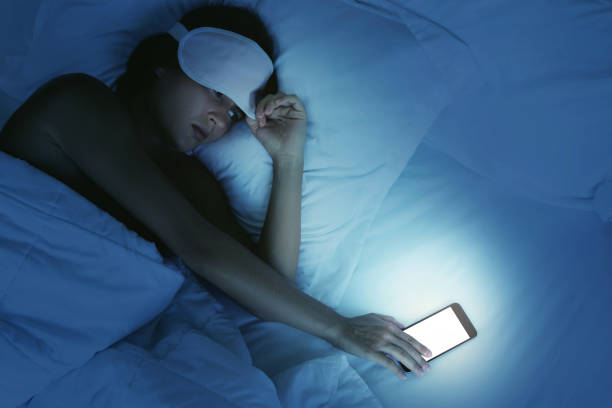 In this image, a woman is laying in her bed wearing an eye mask. She is peaking under her eye mask as she reaches for her phone that is lit up.