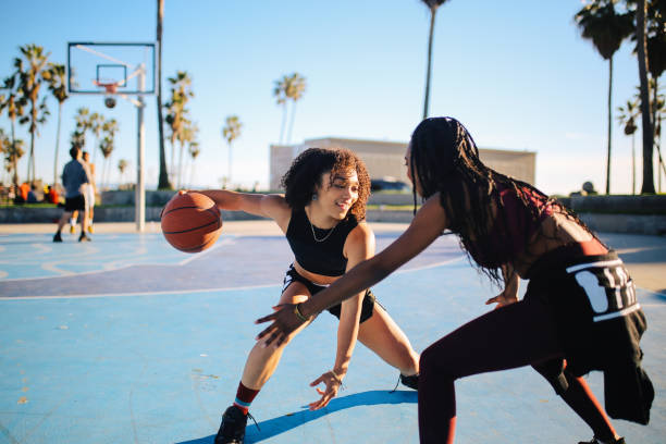 Two young African American woman are by the beach, playing basketball on a basketball court. One of the women has the ball in her hand as the other one tries defending.