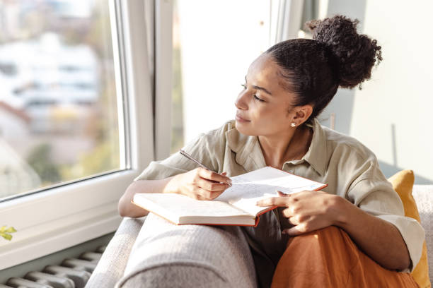 A young African American woman is sitting at her home on the couch as she looks out the window. She is writing in a notebook that is opened in front of her.