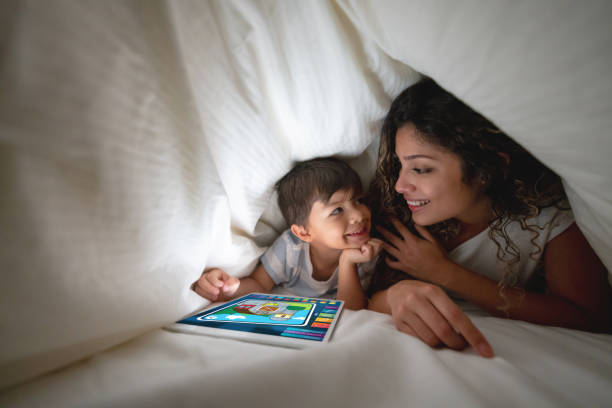 In this image, a mother and son are playing together under the bed covers. They are both smiling at each other as the son plays on the tablet in front of him.