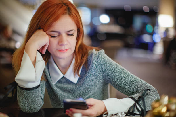 In this image, a red headed young woman is sitting at a cafe using her phone. One of her hands is holding her head up and she uses her phone with her other hand.