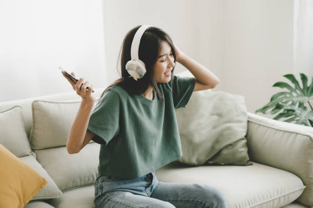 An Asian American young woman is sitting in her home on her couch. She is listening to music on her phone with a headset on her head.