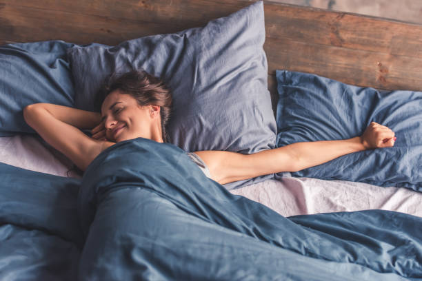 A young woman is laying in her bed in the morning, stretching her arms and smiling.