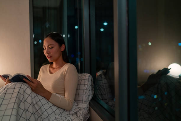 In this image, an Asian American woman is sitting on her bed by the window reading a novel. She is covered by her blanket.