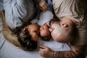 A married couple is laying in their bed with their newborn baby in between them. The parents are kissing the baby on both sides of the babies cheek.