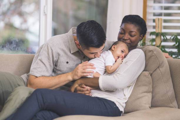 A biracial couple is sitting in their home on their couch. The African American mother is holding the child on her chest, as the father kisses the baby on it's back.