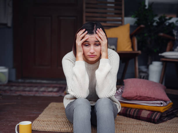 A young Asian American woman is sitting on the front porch of her house. She has blankets and pillows beside her on the floor. Her yellow coffee mug is beside her. She is holding her head up with her hands as her elbows rest on her knees.