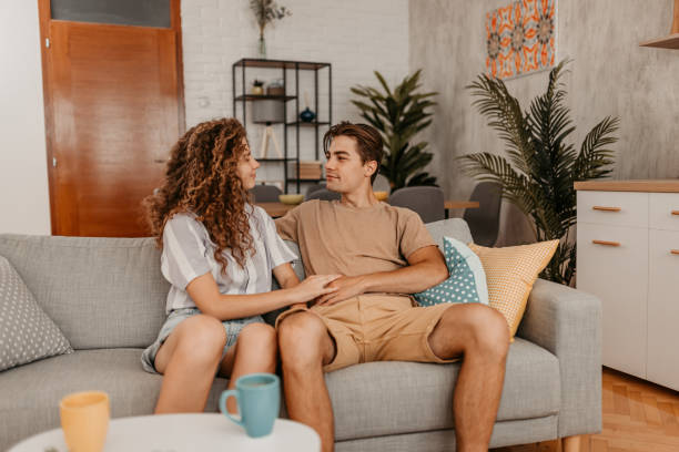 A young couple is sitting in their living room on the couch beside each other. They are gazing at each others eyes as they smile at one another.
