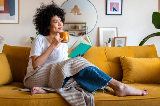 A young African American woman is sitting in her home on the couch. She has her legs up on the couch as she has a blanket draped on her. She is holding a coffee mug in one hand and an open book in her other hand.