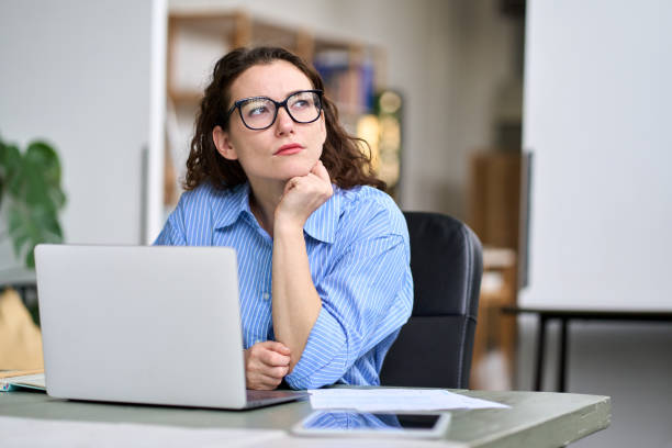 A young beautiful American woman is sitting at her desk in her office. She has her laptop opened in front of her. She is wearing glasses and is resting her head on her hand.