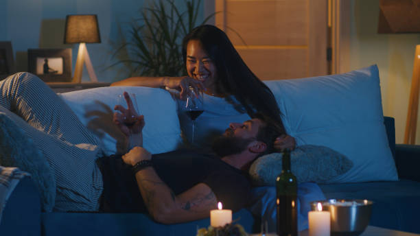 A biracial couple is in their living room on the couch. The lights are dimmed and there are candles lit on the coffee table. The man is laying on his partner's lap as they smile and laugh.