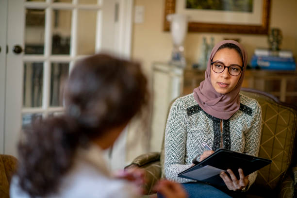 A young Arab therapist is sitting in her office during a therapy session. The therapist is wearing a hijab on her head, along with her glasses. In her hands she is holding a notepad and pen as her female client sits across from her.