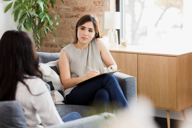 A young woman is sitting in an office across from her therapist in a therapy session. The therapist has a clipboard on her lap writing notes as the client speaks to her.