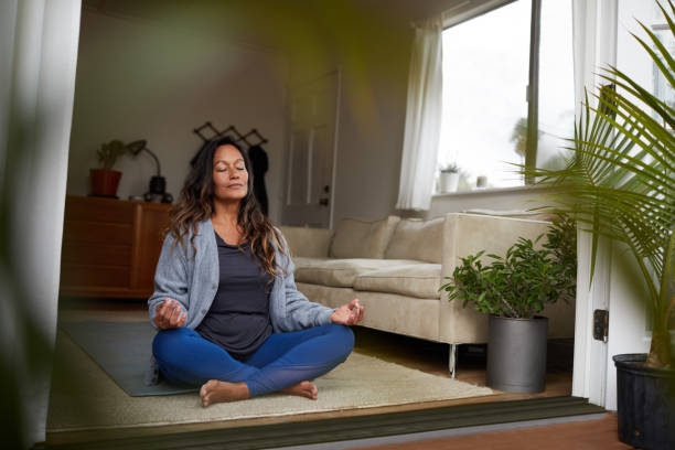 A young woman is sitting on the floor of her living room facing the outside with the doors open. She is sitting in a yoga position with her hands on her knees and her eyes closed.
