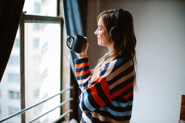 A young woman is standing in her studio apartment as she looks outside of the window. She has headphones on her head and is holding a black coffee mug.