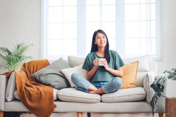 A young Asian American woman is sitting in her home on the couch. She has her legs crossed on the couch as she holds a coffee mug in her hands. She is smiling and looking up.