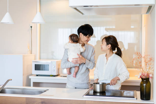 An Asian American couple is standing in their kitchen cooking dinner. The husband is holding an infant child in his arms standing beside his wife. The wife is cooking a pot on the stove.