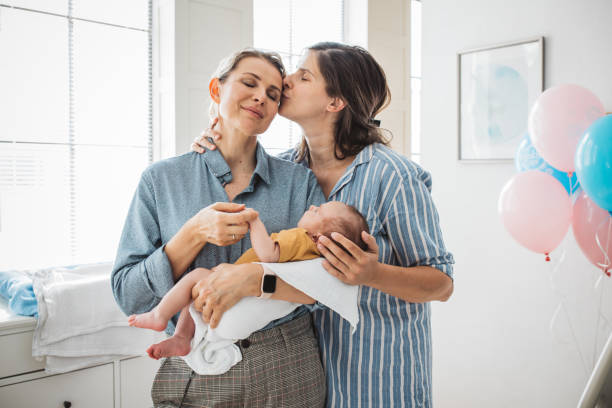 An LGBT couple is celebrating the birth of their infant child. The two women are standing beside each other as one of the carries the child and the other is kissing the other woman on the cheek.