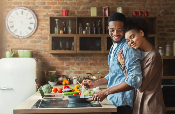 A young African American couple is standing in their kitchen. The husband is cooking food on the stove as his wife embraces him from behind him.