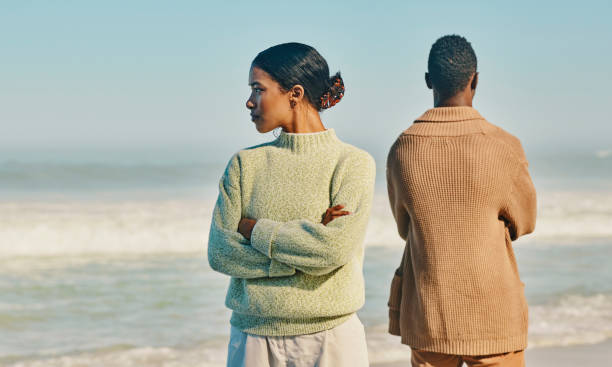 An African American couple is standing at the beach, facing their backs to one another. Both of their arms are crossed and they are looking away from each other.