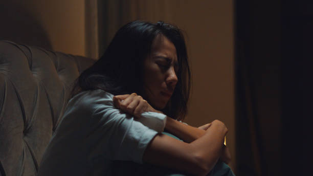 An Asian American woman is sitting in a dark room with her arms wrapped around herself. She is holding a tissue in one hand as she has a distressed facial expression.