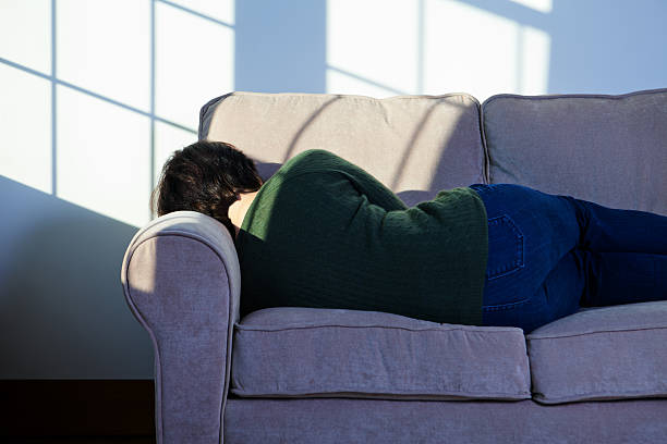 A woman is laying on the couch in her living room. She is facing her back towards the camera as she lays her head on the armrest of the couch.