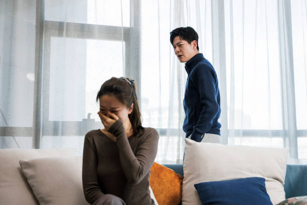 A young Asian couple is in the midst of a fight as the woman sits on the couch and the man stands behind her. She has her hand covering her mouth as she cries.