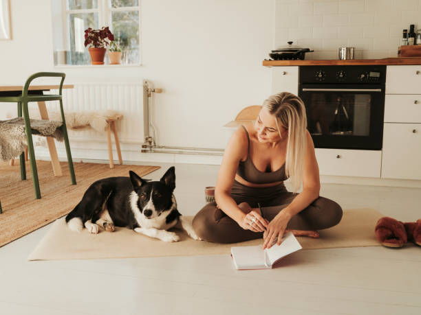 A young blonde woman is sitting on a yoga mat in her home. Her dog is laying beside her as she writes in her journal.