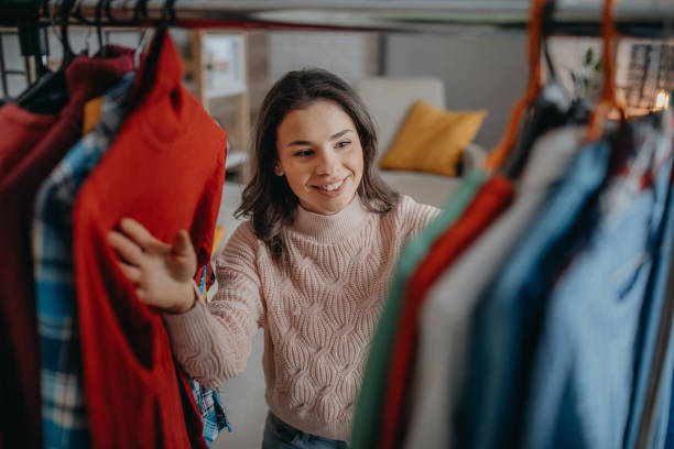 a beautiful young woman is in her closet looking through a rack of clothing. There are colorful clothes hanging as she searches through them and smiles.