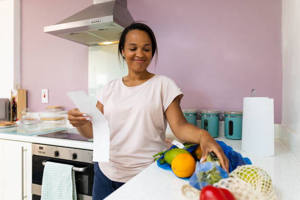 A young black woman is standing in her kitchen looking at the groceries that are on the countertop. She is holding a long grocery list.