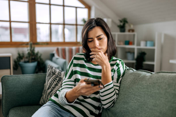 A young woman is sitting on the couch and holding her phone in her hand. She has her hand covering her mouth as she looks at something on her phone.